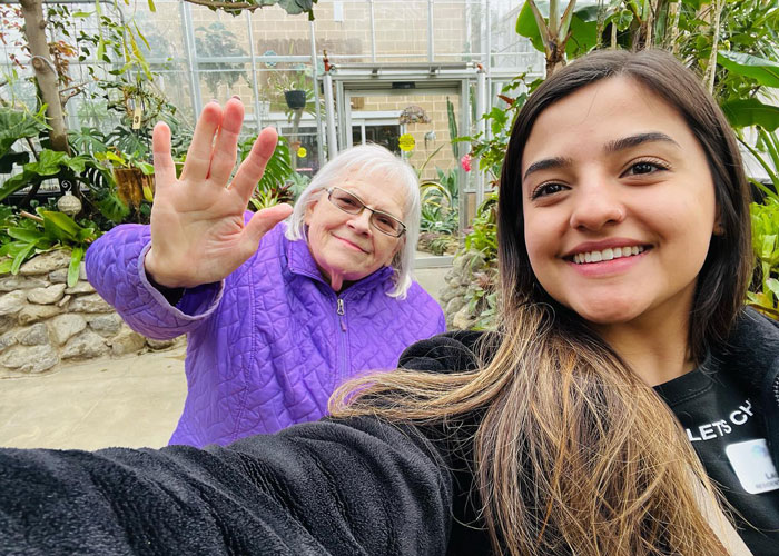 Elderly woman in a purple jacket waves cheerfully while taking a selfie with a smiling young woman during an outing at a greenhouse.