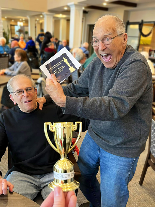 Two elderly men celebrating a victory, with one holding a 'People's Choice' trophy and the other excitedly showing an award certificate, surrounded by a joyful community.