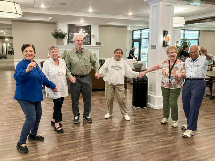 A group of elderly individuals holding hands and dancing joyfully in a spacious and well-lit community area.