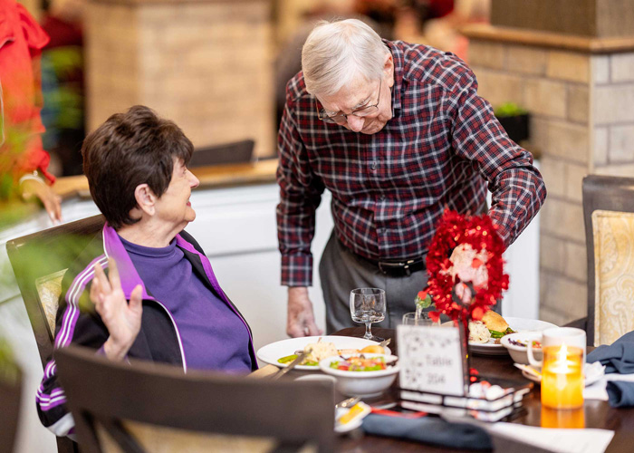 Elderly man leans over to talk to an elderly woman sitting at a dining table, both smiling and enjoying a meal together.