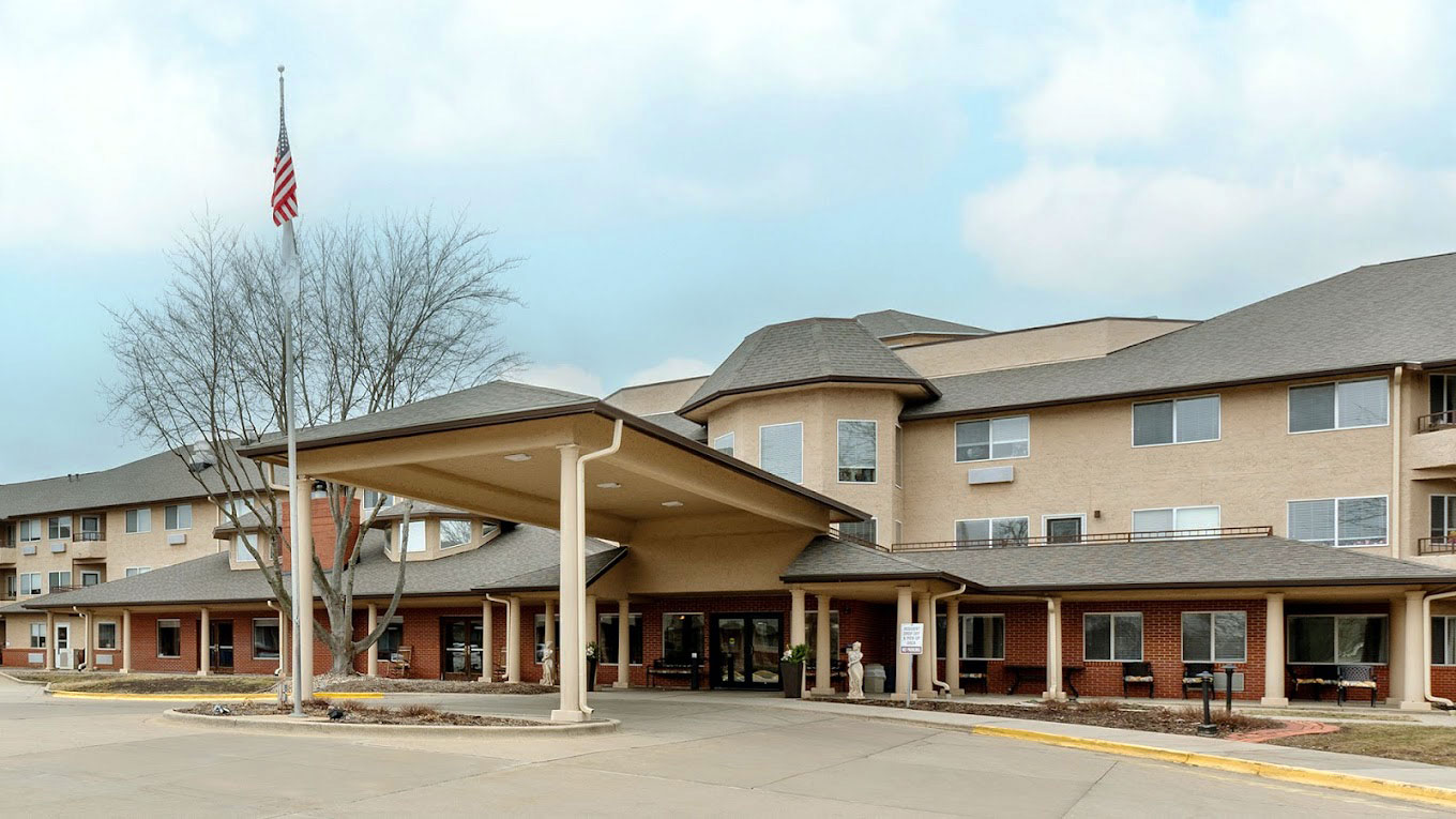 The Montvale Senior Living community features a welcoming exterior with a covered entrance, well-maintained landscaping, and a prominent American flag. The building's design combines modern amenities with a homely atmosphere, providing a comfortable and inviting environment for its residents.