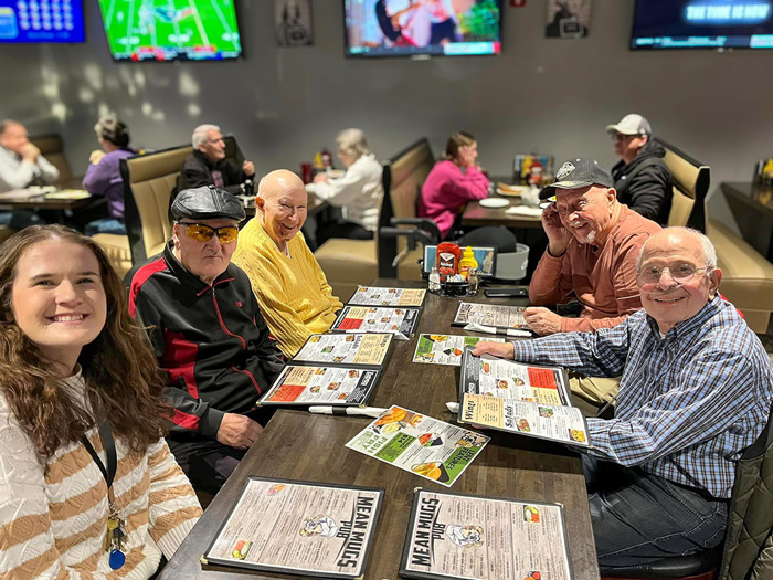 Senior residents enjoying a meal together at a sports-themed restaurant, with menus and TVs in the background.