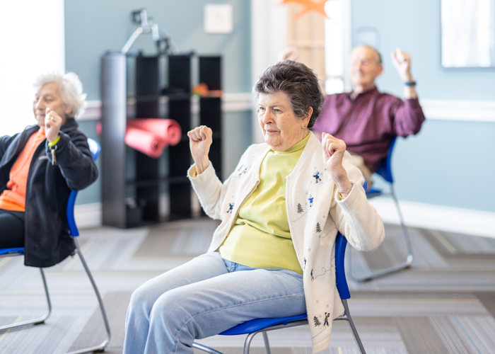 Elderly woman in a yellow shirt and white cardigan participating in a seated exercise class, with other seniors actively joining in the background.