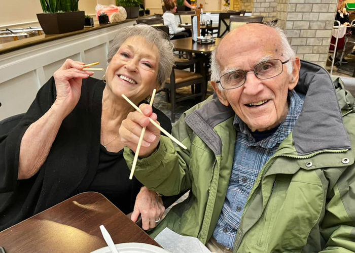 Two senior residents smile while holding chopsticks during a meal at a senior living community, enjoying a shared dining experience together.