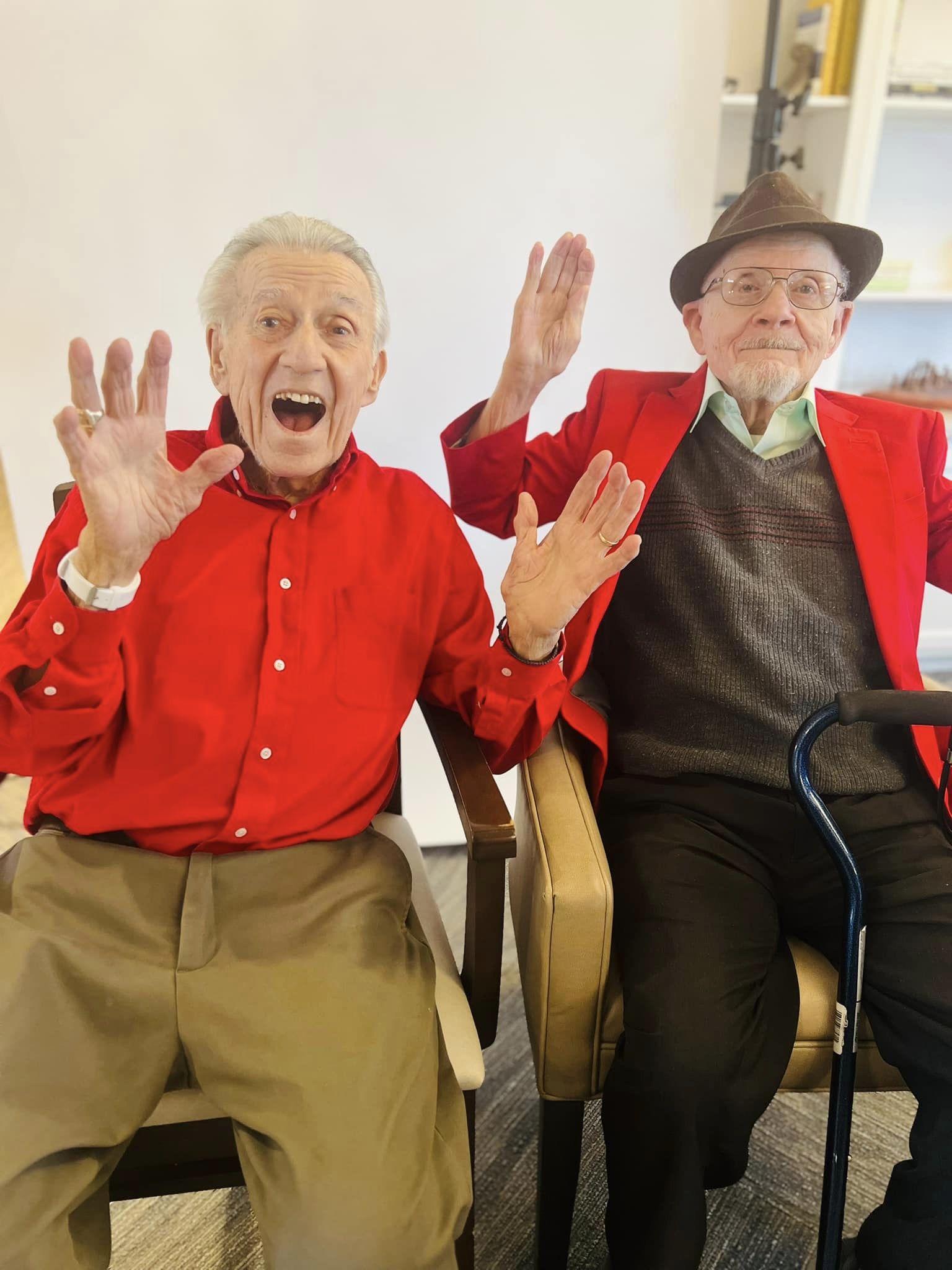 Two senior residents, both dressed in red jackets, smile and raise their hands in a playful gesture while sitting together at a senior living community event.