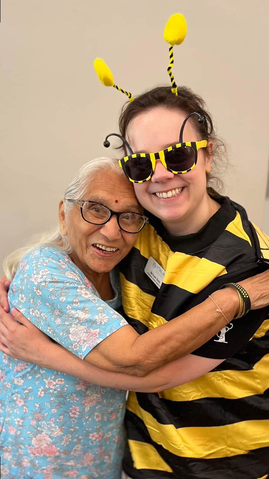 Senior resident in a floral dress hugs a smiling team member dressed in a bee costume with yellow antennae and glasses, creating a heartwarming moment at a community event.