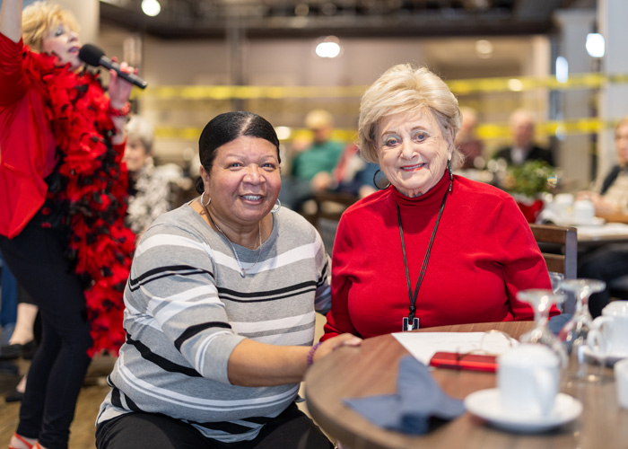 Two senior residents sit together at a table during an event in a senior living community, smiling as they enjoy the entertainment provided by a singer in the background.