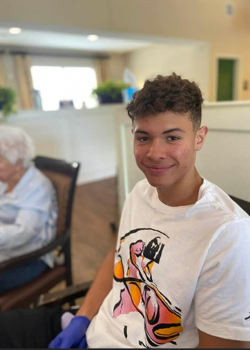 A young staff member smiles while seated at a senior living community, wearing a colorful graphic t-shirt, engaging with residents in a relaxed and welcoming environment.