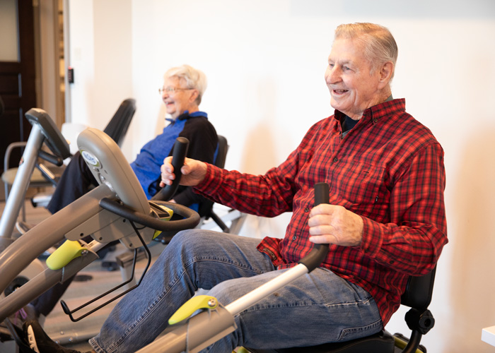 Two elderly individuals, a man in a red plaid shirt and a woman in a blue jacket, smile while exercising on recumbent bikes in a bright and welcoming fitness room.