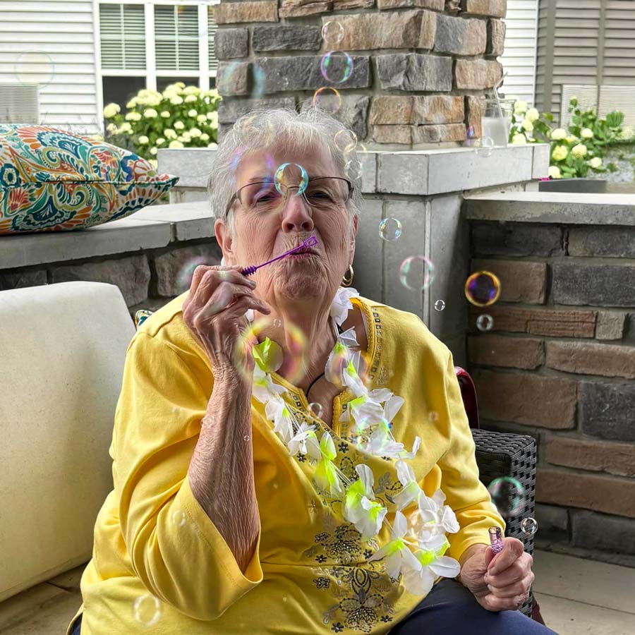 Smiling senior resident wearing a yellow shirt and a flower lei blows bubbles while sitting on a porch, enjoying a playful and joyful moment outdoors.