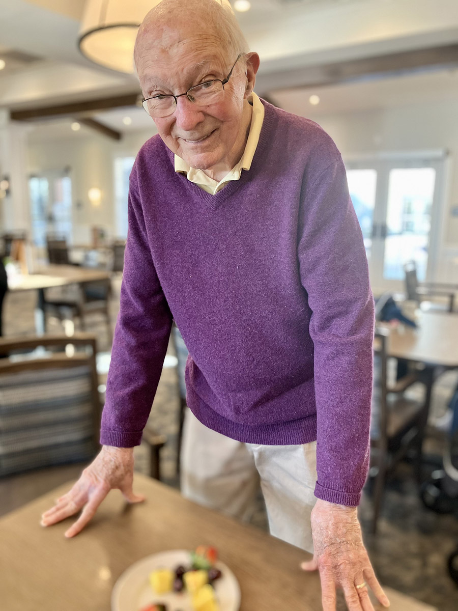 A senior resident wearing a purple sweater smiles warmly while standing at a table with a plate of fresh fruit in a bright and welcoming dining area at Vitalia Senior Living.