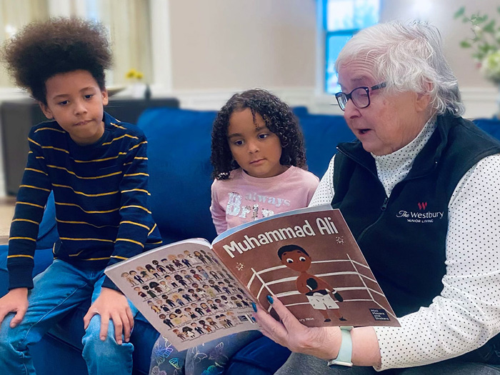 A senior resident wearing a vest with 'The Westbury Senior Living' logo reads a 'Muhammad Ali' children's book to two attentive young children, sitting together on a couch.