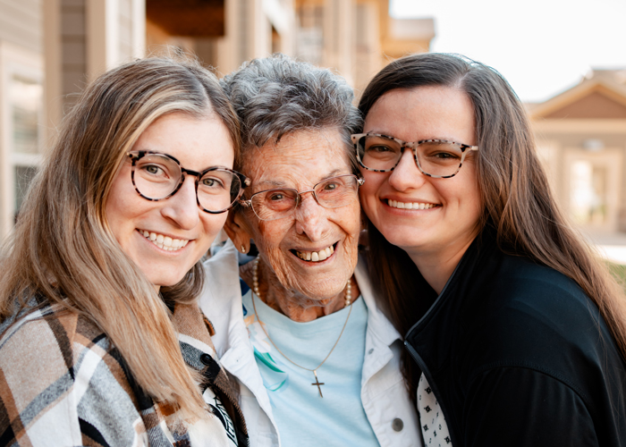 A senior resident smiles between two younger women, all wearing glasses, sharing a happy moment outdoors at a senior living community.