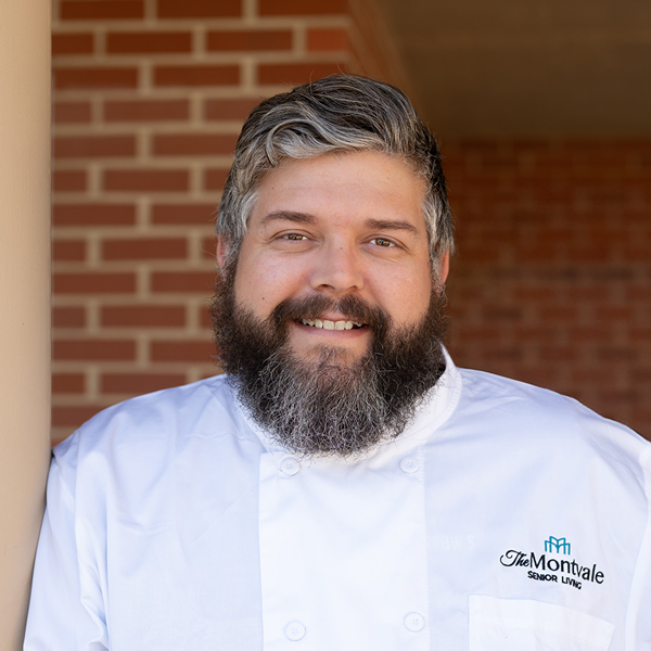 Cody Clinton, Culinary Director at Montvale Senior Living, smiling in a white chef's coat embroidered with the community logo, standing in front of a brick wall.