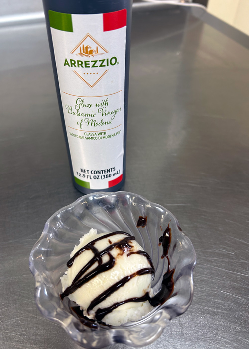 Vanilla ice cream scoop drizzled with balsamic glaze in a glass dish, with an Arrezzio balsamic vinegar bottle in the background on a stainless steel counter.