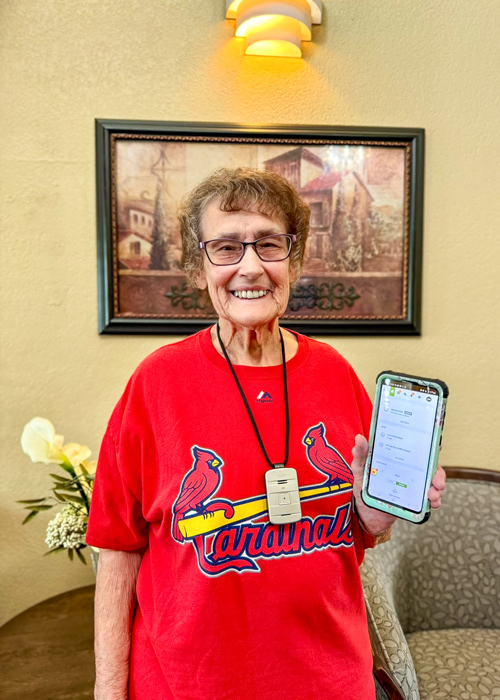 Senior resident wearing a red Cardinals shirt smiles while holding up a smartphone displaying the Arrow app in a warmly decorated room.
