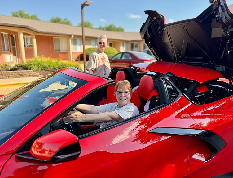 A senior woman smiles excitedly from the driver's seat of a bright red sports car.
