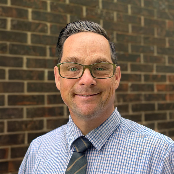 Aaron Mazrim, Plant Operations Director at Montvale Senior Living, smiling in a professional headshot. He has short dark hair, wears glasses, a blue checkered dress shirt, and a striped tie, standing in front of a brown brick wall background.