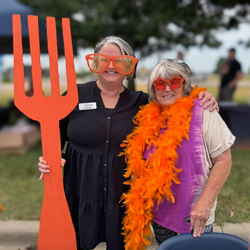 Two women smile at an outdoor event, one wearing a large, orange feather boa and the other holding a large, orange prop fork.