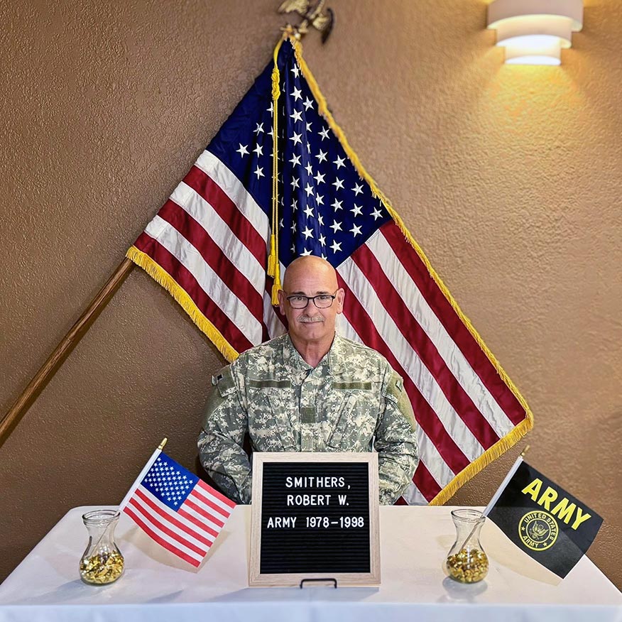 An army veteran in uniform smiles in front of a flag on Veterans Day.