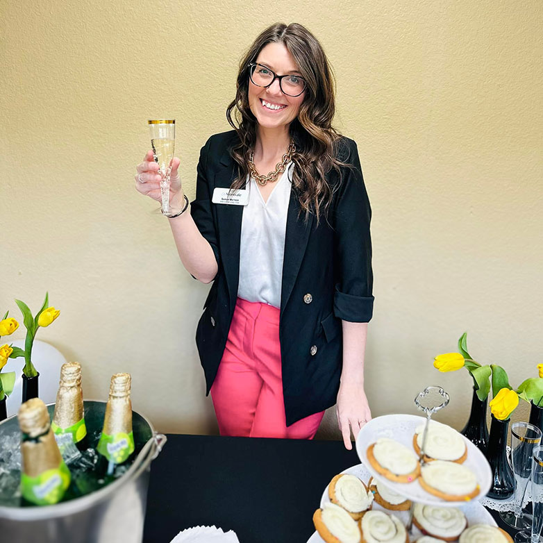 A team member in business dress smiles while raising a drink near a table full of elegant treats.