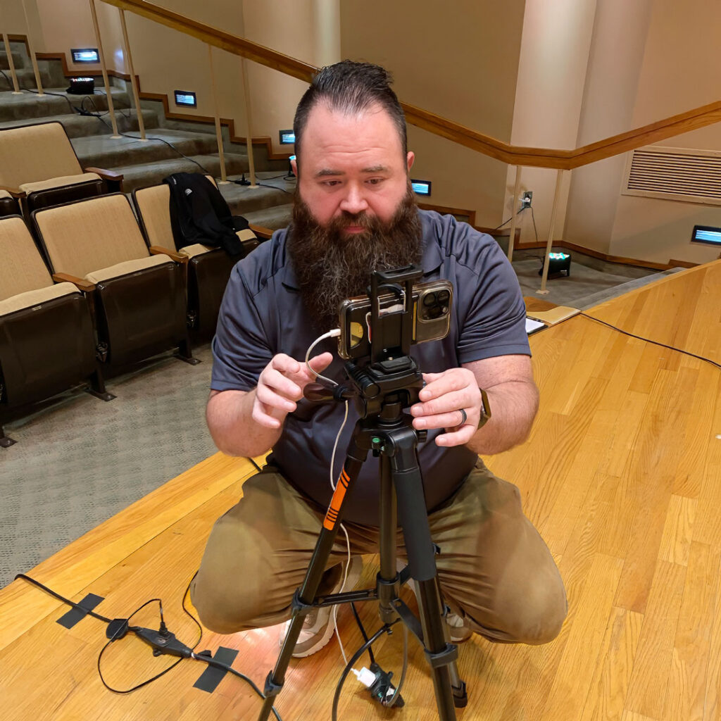 Colby Boyes, Arrow Senior Living Technical Support Specialist, adjusts a smartphone camera on a tripod during a Thoughtful Connections podcast recording.