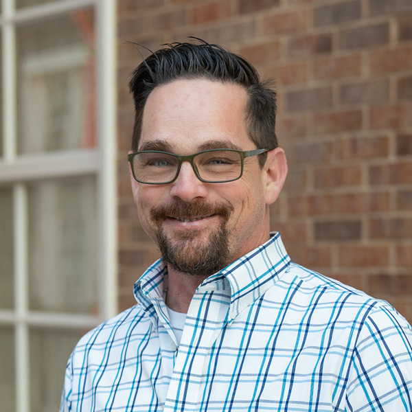 Aaron Mazrim, Plant Operations Director at Montvale Senior Living, smiling in a professional outdoor headshot with dark styled hair, glasses, a goatee, a blue plaid button-up shirt, and a softly blurred brick background.