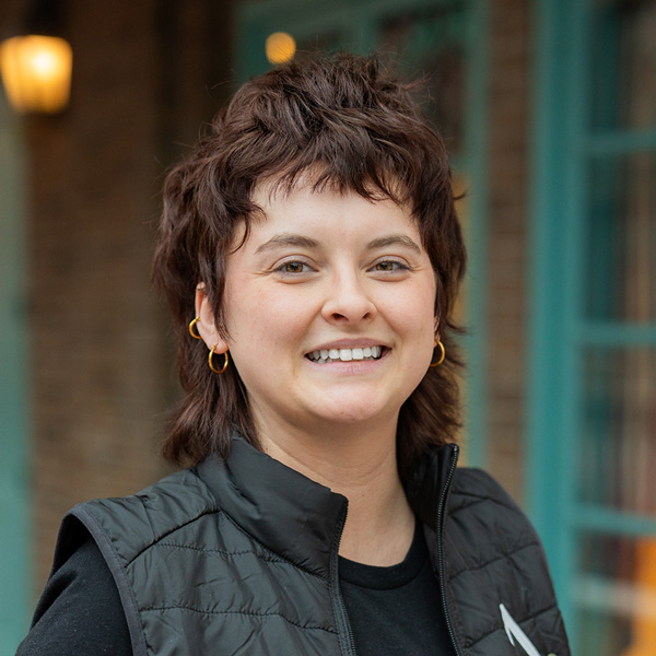 Jane Sorenson, Resident Services Director at Montvale Senior Living, smiling in a professional outdoor headshot with short textured dark hair, gold hoop earrings, a black vest, and softly blurred brick and teal window details in the background.