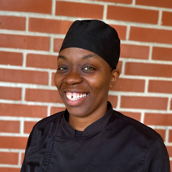Carolyn “Jade” Henderson, Culinary Director at Montvale Senior Living, smiling in a professional headshot wearing a black chef coat and black head covering against a red brick wall background.